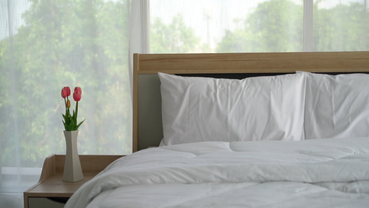 Uređen krevet sa belom posteljinom i drvenim uzglavljem pored malog stočića sa cvećem